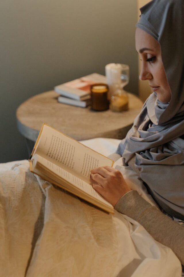 A woman in traditional Islamic attire reads a book in a cozy bedroom with a candle-lit ambiance.