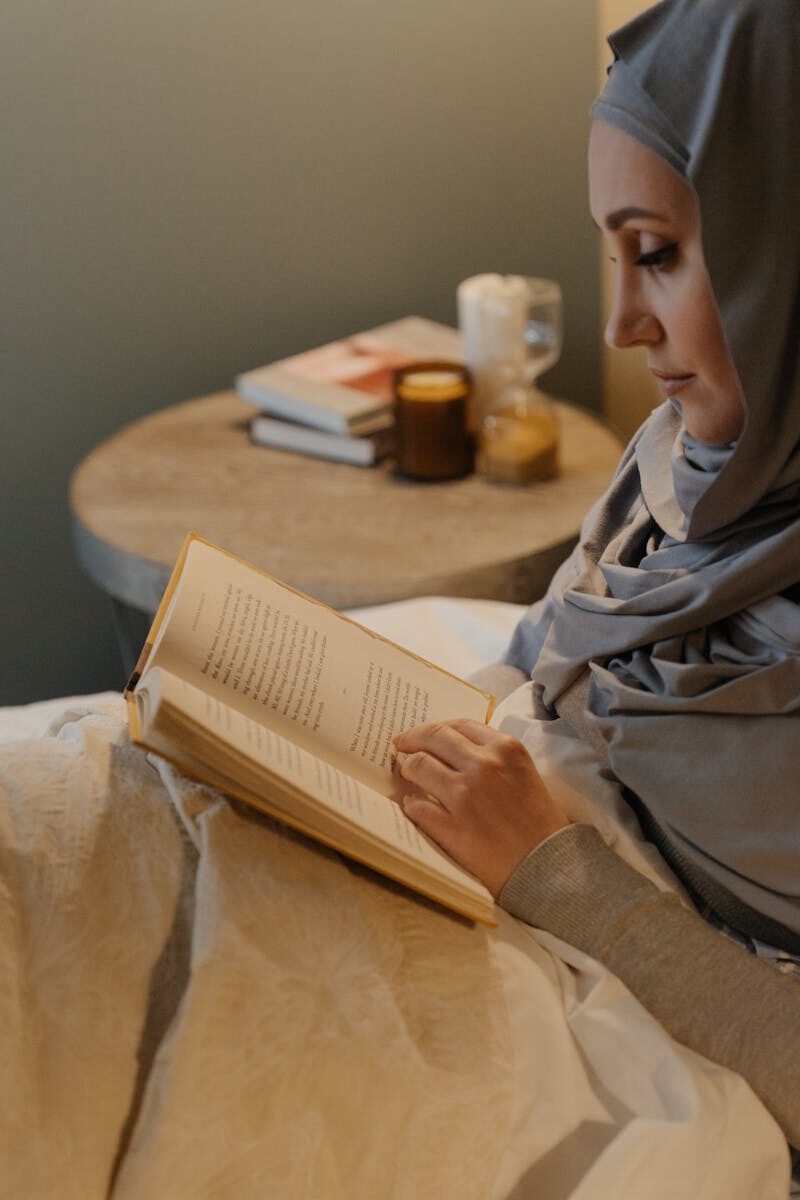 A woman in traditional Islamic attire reads a book in a cozy bedroom with a candle-lit ambiance.