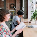 Two individuals reading at a wooden table in a modern urban cafe setting with large windows.