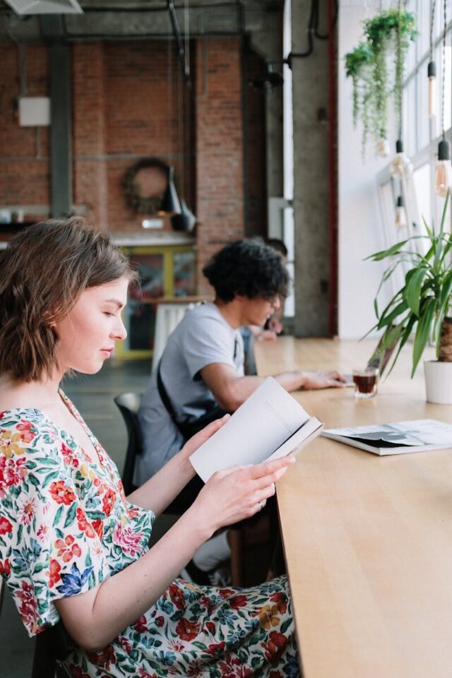 Two individuals reading at a wooden table in a modern urban cafe setting with large windows.
