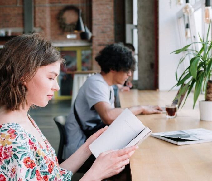 Two individuals reading at a wooden table in a modern urban cafe setting with large windows.