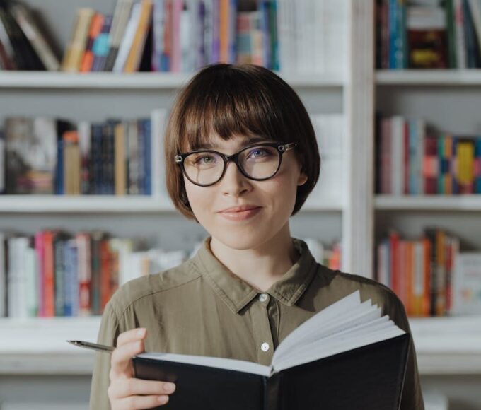 Smiling woman in a library holding a book, surrounded by bookshelves.