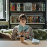 A young woman enjoys reading in a cozy library, holding a book with a coffee on the table.