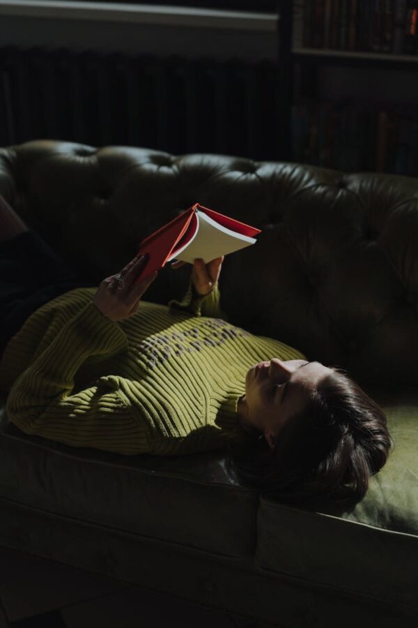 A young woman lies on a green sofa reading a red book, enjoying leisure indoors.