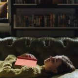 A young woman in a green sweater naps with a book in a cozy library setting.