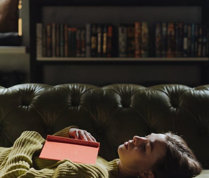 A young woman in a green sweater naps with a book in a cozy library setting.