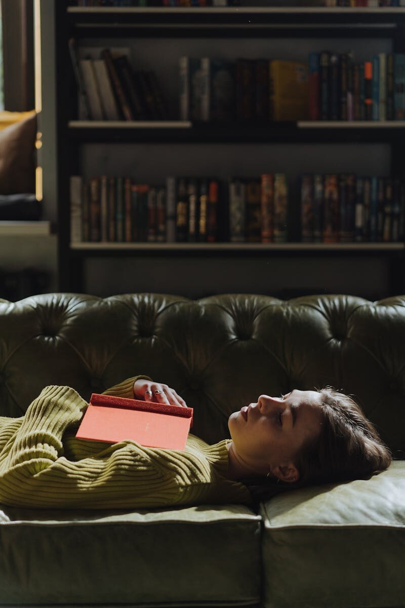 A young woman in a green sweater naps with a book in a cozy library setting.