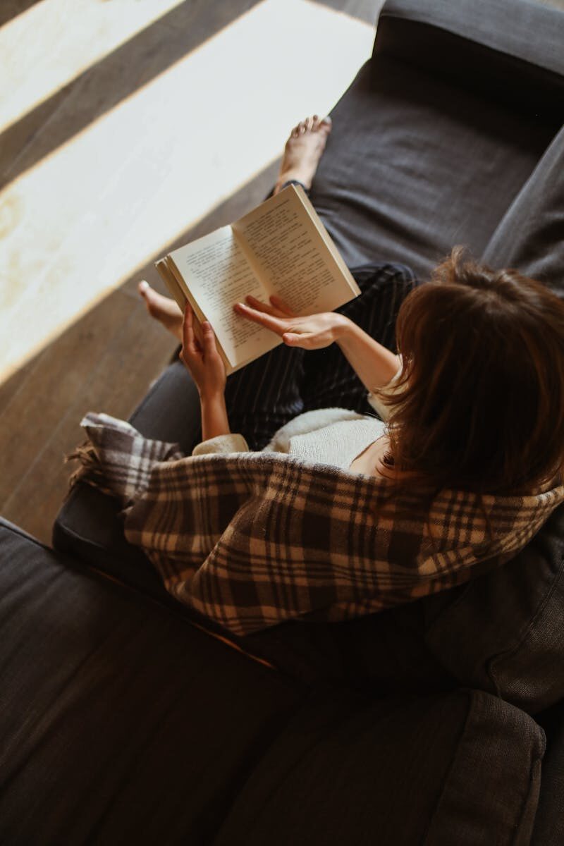 Woman relaxing with a book on a cozy sofa bathed in sunlight.