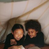 Two children happily reading under a cozy blanket fort with warm lighting.
