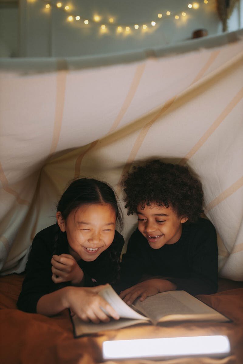 Two children happily reading under a cozy blanket fort with warm lighting.