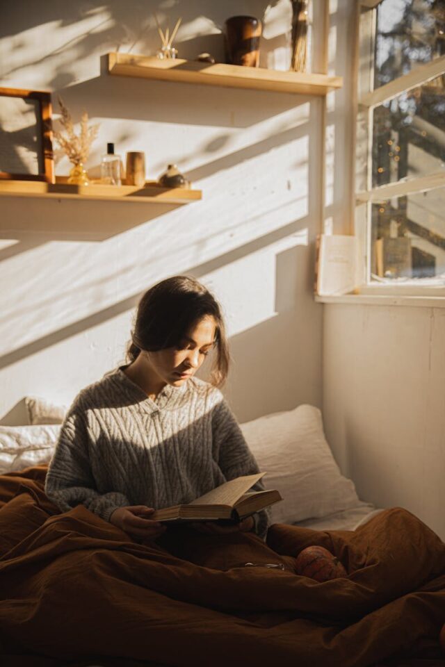 Woman enjoying a peaceful reading moment in a sunlit bedroom, cozy atmosphere.