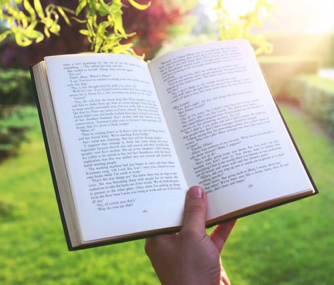 Open book held by hand in a bright green garden under sunlight, evoking relaxation and leisure.