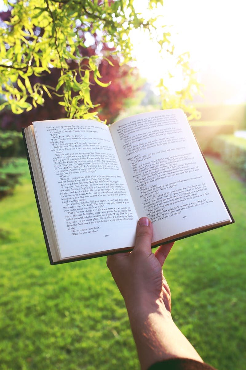 Open book held by hand in a bright green garden under sunlight, evoking relaxation and leisure.