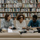 Three students engaged in group study session surrounded by bookshelves.