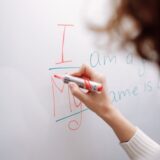 A hand with a marker writing on a whiteboard indoors. Educational context.