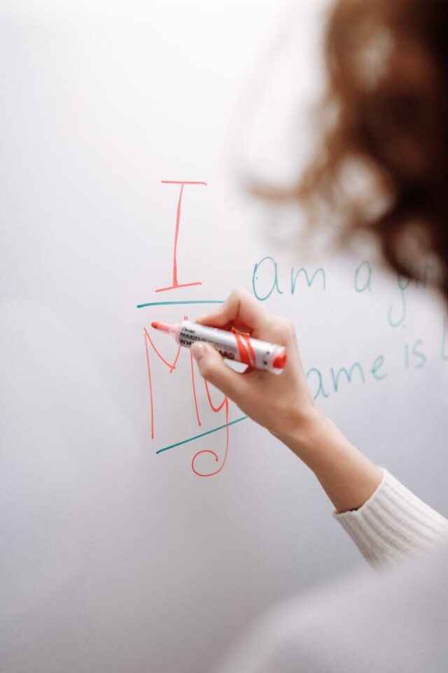 A hand with a marker writing on a whiteboard indoors. Educational context.