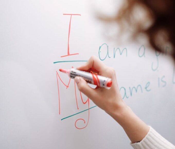 A hand with a marker writing on a whiteboard indoors. Educational context.