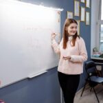 Young woman teaching English lesson in a classroom, writing on a whiteboard.