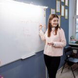 Young woman teaching English lesson in a classroom, writing on a whiteboard.