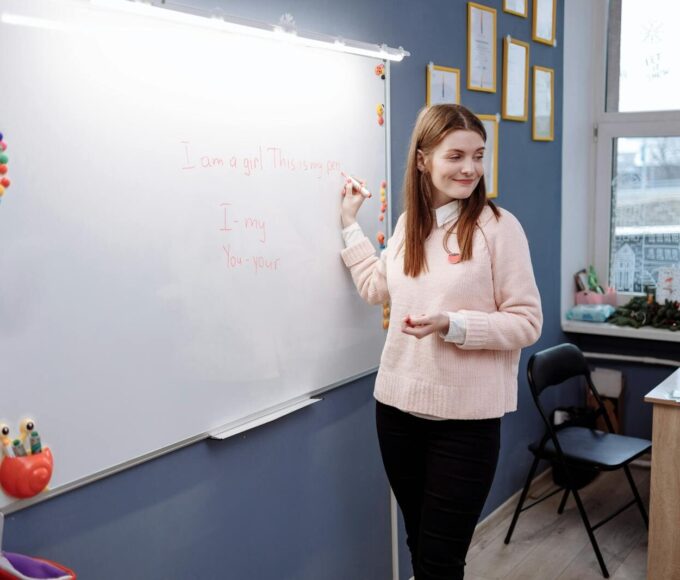 Young woman teaching English lesson in a classroom, writing on a whiteboard.