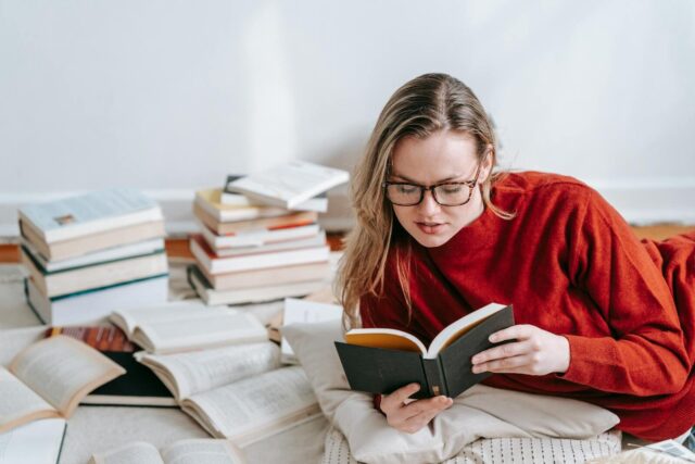 A woman in a red sweater reading a book on the floor with stacks of books around her indoors.