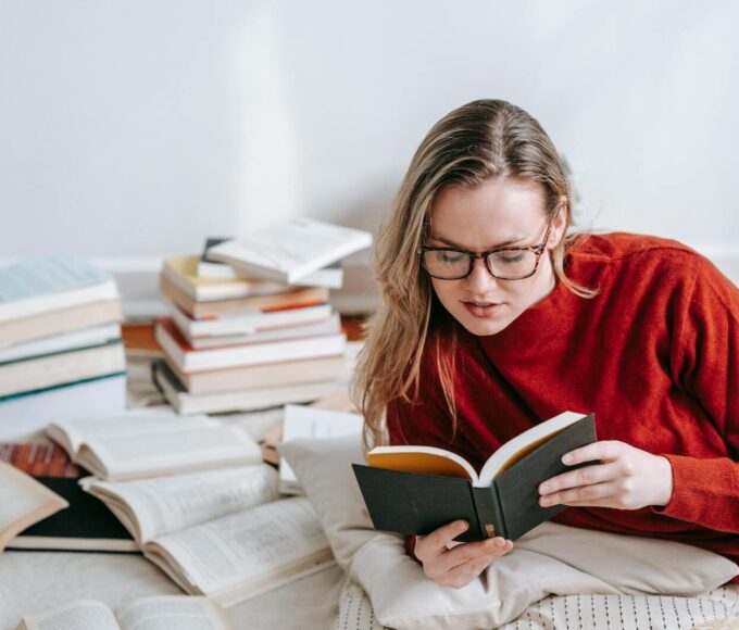 A woman in a red sweater reading a book on the floor with stacks of books around her indoors.