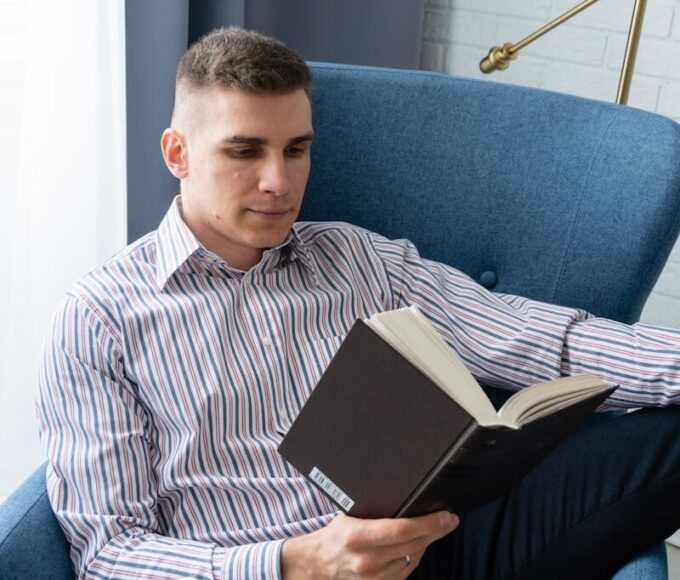 Adult man reading in an armchair indoors, showcasing relaxation and leisure.