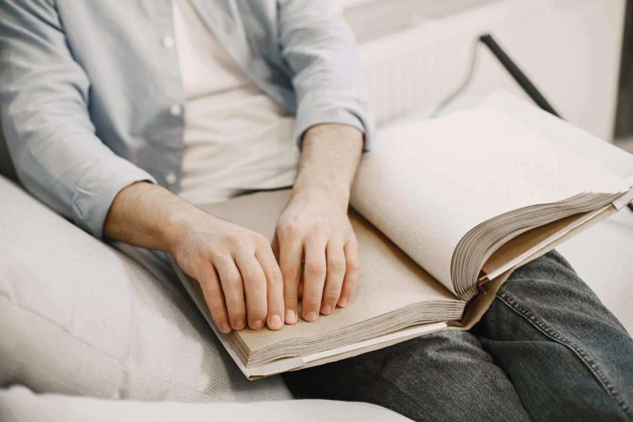 An adult reading a braille book indoors, highlighting visual impairment awareness.