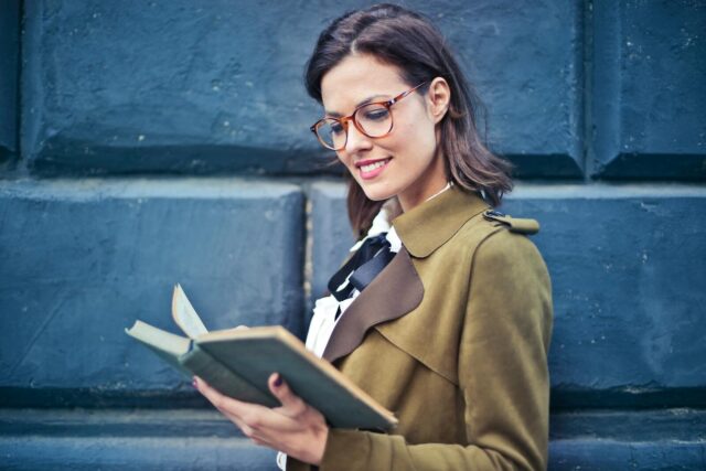 Smiling woman enjoying a book while standing against a textured blue wall.
