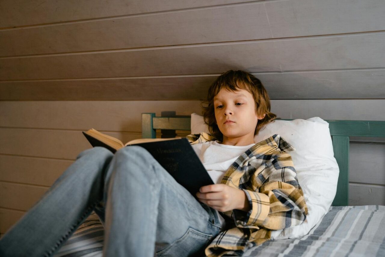 A young boy is comfortably reading a book while lying in bed, creating a serene indoor atmosphere.