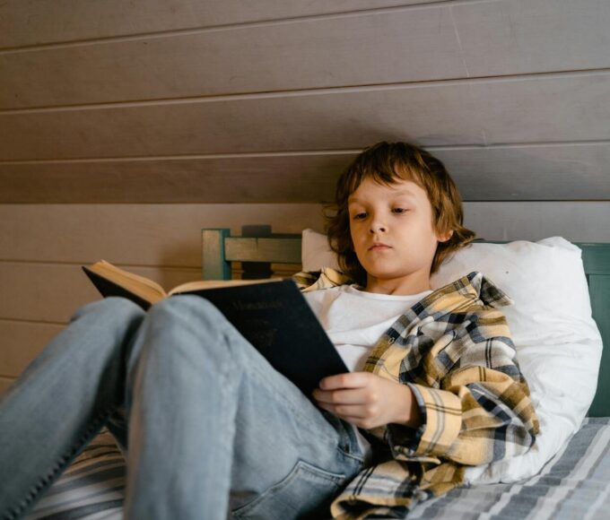 A young boy is comfortably reading a book while lying in bed, creating a serene indoor atmosphere.