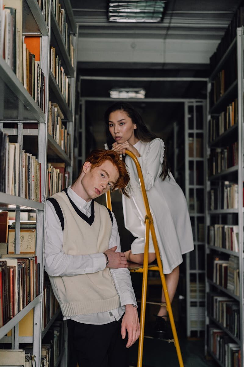 Fashionable young adults posing on a ladder among library bookshelves.