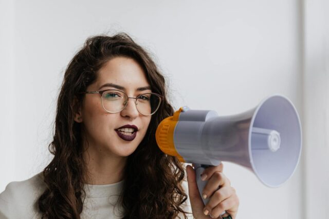 Confident woman with eyeglasses speaking into a megaphone in a bright room.