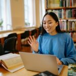 Smiling student using laptop in a library for studying and video calls.