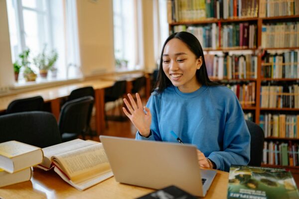 Smiling student using laptop in a library for studying and video calls.