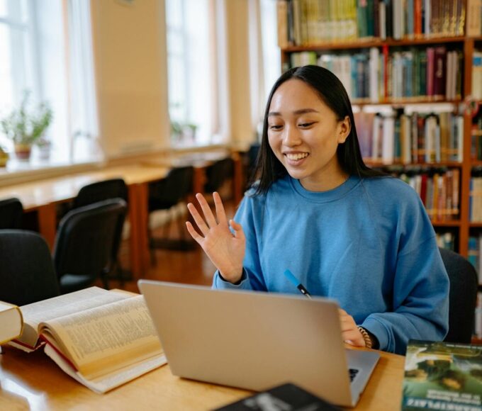 Smiling student using laptop in a library for studying and video calls.
