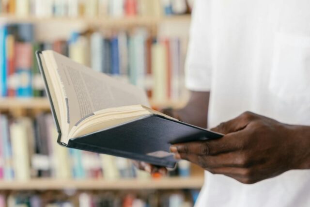 A person reads a book in a library, focusing on hands holding an open book.