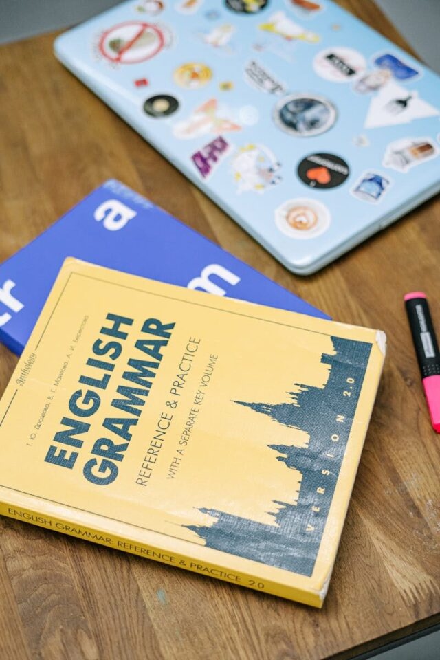 High-angle view of an English grammar book and a laptop with stickers on a wooden desk.