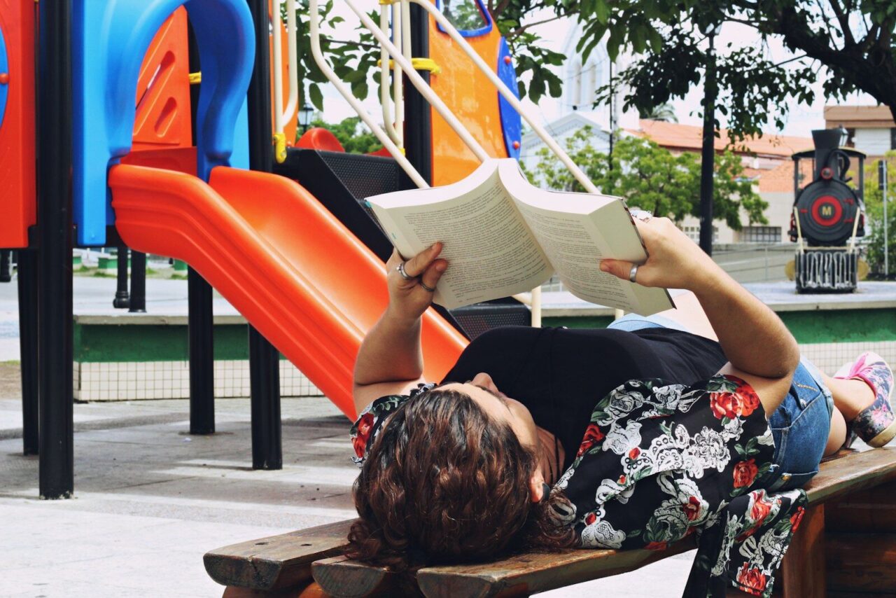 woman lying on bench reading book at playground during daytime