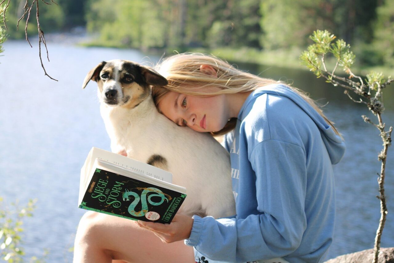 Girl and dog enjoy a book near a lake.