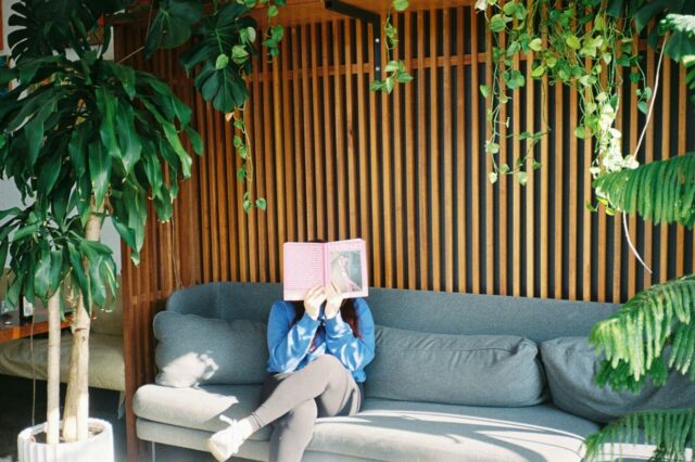 Person reads a book amidst plants on a cozy couch.