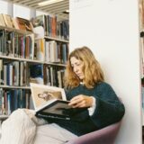 Woman reads a book surrounded by bookshelves.